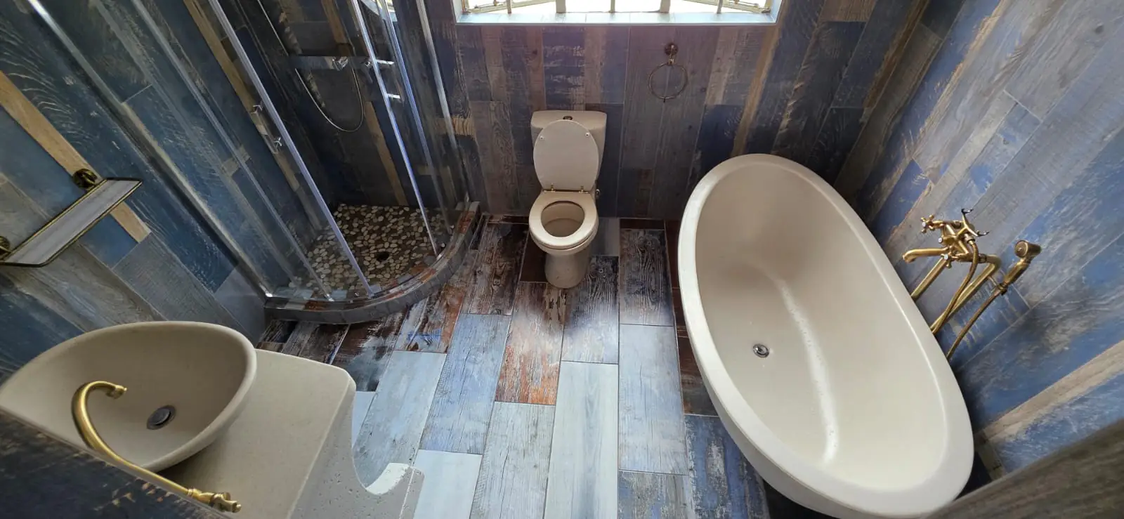 The rebuilt bathroom clad in blue and grey wood-look plank tiles, with a freestanding oval bath, brushed brass mixer and hand shower, a curved glass shower enclosure over a pebble floor and a cast vanity holding a stone vessel basin.