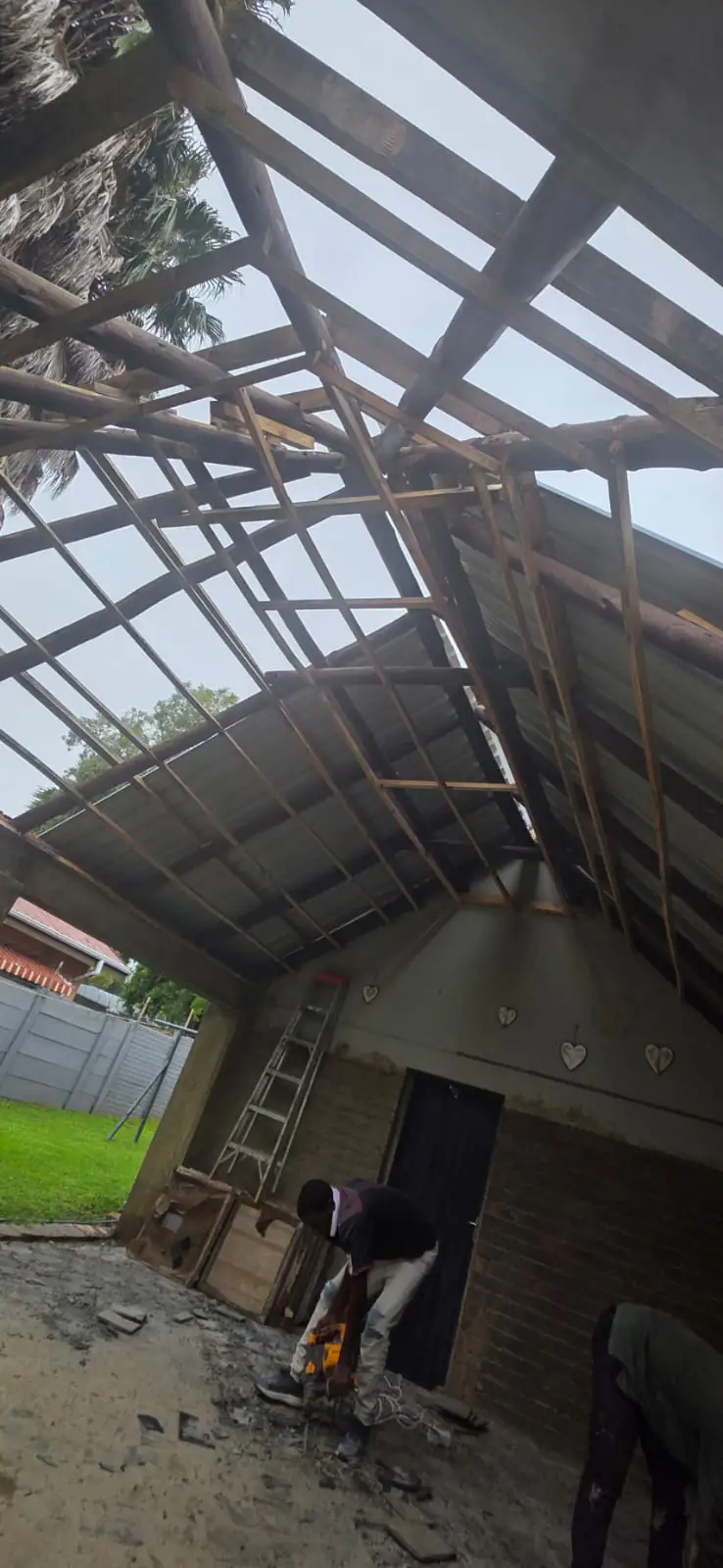 Roof timbers stripped back to bare purlins and rafters over an unfinished patio, with a worker cutting timber below and a ladder against the gable wall.